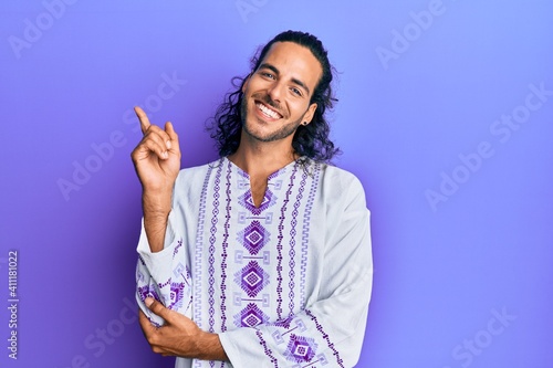 Young handsome man with long hair wearing bohemian and hippie shirt with a big smile on face, pointing with hand and finger to the side looking at the camera. © Krakenimages.com