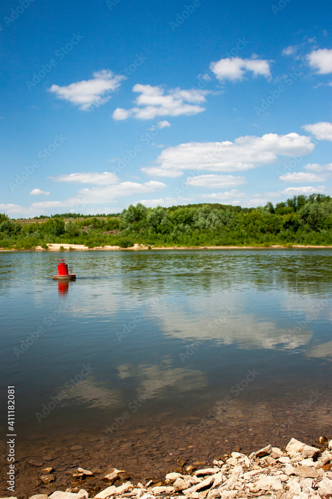 View from the river bank to the red buoy on a bright summer day. The red buoy is reflected in the calm water of the river. Blue sky and clouds are reflected in the river.