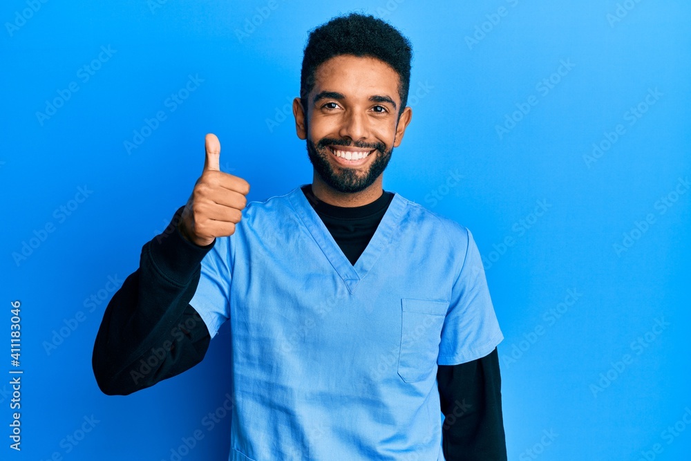 Fototapeta premium Handsome hispanic man with beard wearing blue male nurse uniform smiling happy and positive, thumb up doing excellent and approval sign