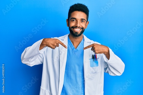 Handsome hispanic man with beard wearing doctor uniform looking confident with smile on face, pointing oneself with fingers proud and happy.