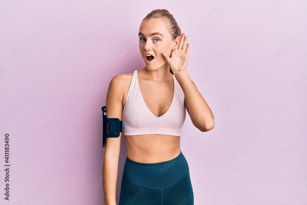 Beautiful blonde woman wearing sportswear and arm band smiling with hand over ear listening an hearing to rumor or gossip. deafness concept.