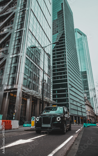 Fotografi London UK January 2021 Vertical shot of a modern hybrid london taxi passing by on leadenhall street, massive tall skyscrapers reaching up high in the background