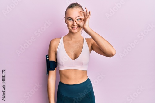 Beautiful blonde woman wearing sportswear and arm band doing ok gesture with hand smiling, eye looking through fingers with happy face. © Krakenimages.com