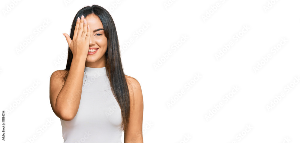Young beautiful hispanic girl wearing casual clothes covering one eye with hand, confident smile on face and surprise emotion. Young beautiful hispanic girl wearing casual clothes covering one eye with hand, confident smile on face and surprise emotion.