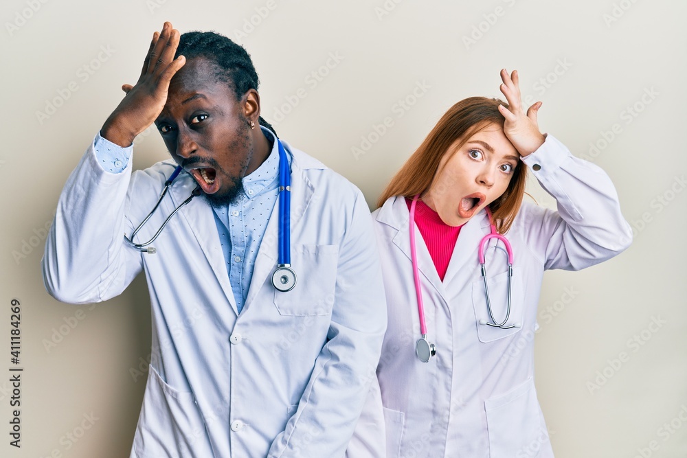 Young interracial couple wearing doctor uniform and stethoscope surprised with hand on head for mistake, remember error. forgot, bad memory concept. Young interracial couple wearing doctor uniform and stethoscope surprised with hand on head for mistake, remember error. forgot, bad memory concept.