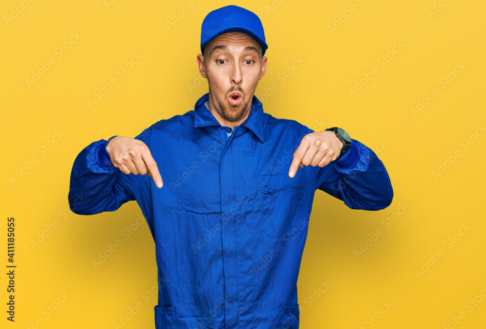 Bald man with beard wearing builder jumpsuit uniform pointing down with fingers showing advertisement, surprised face and open mouth