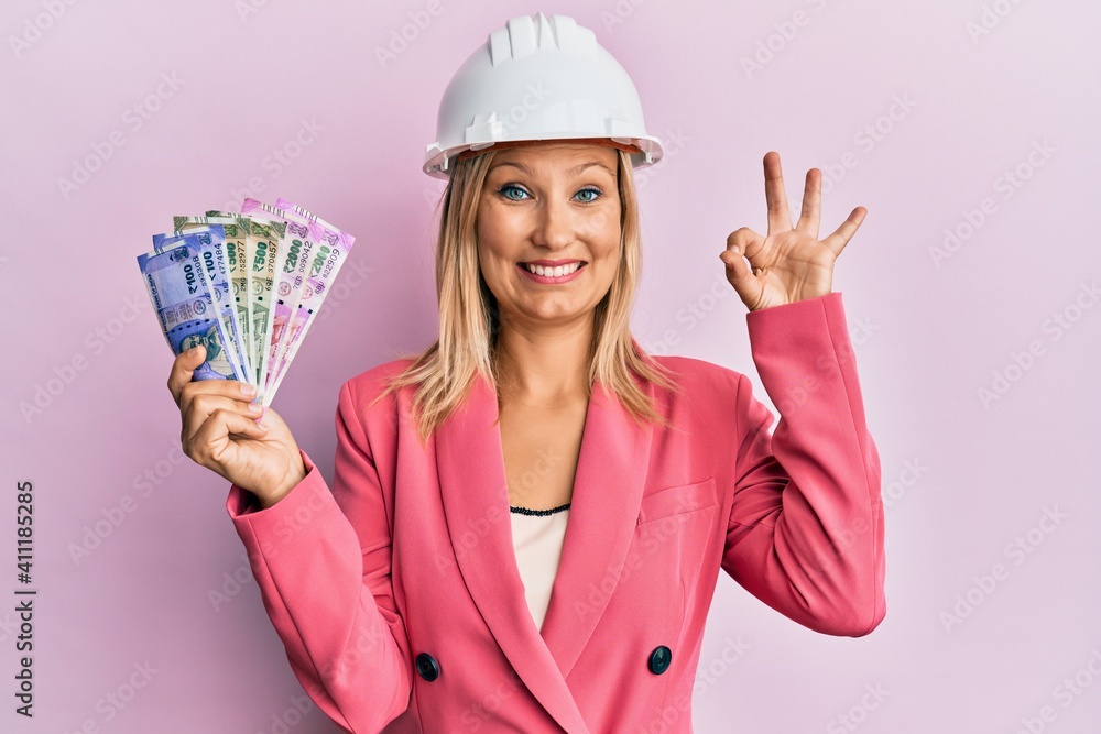 Beautiful middle age blonde woman wearing architect hardhat holding indian rupee doing ok sign with fingers, smiling friendly gesturing excellent symbol Beautiful middle age blonde woman wearing architect hardhat holding indian rupee doing ok sign with fingers, smiling friendly gesturing excellent symbol