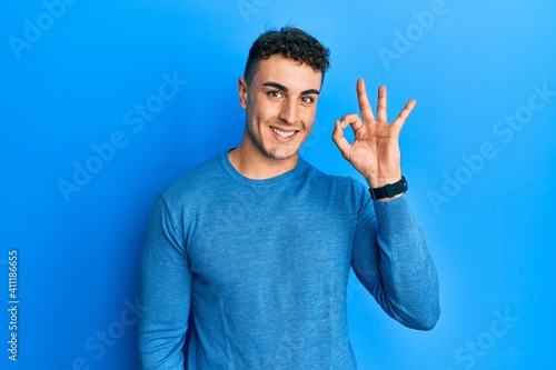 Hispanic young man wearing casual winter sweater smiling positive doing ok sign with hand and fingers. successful expression.