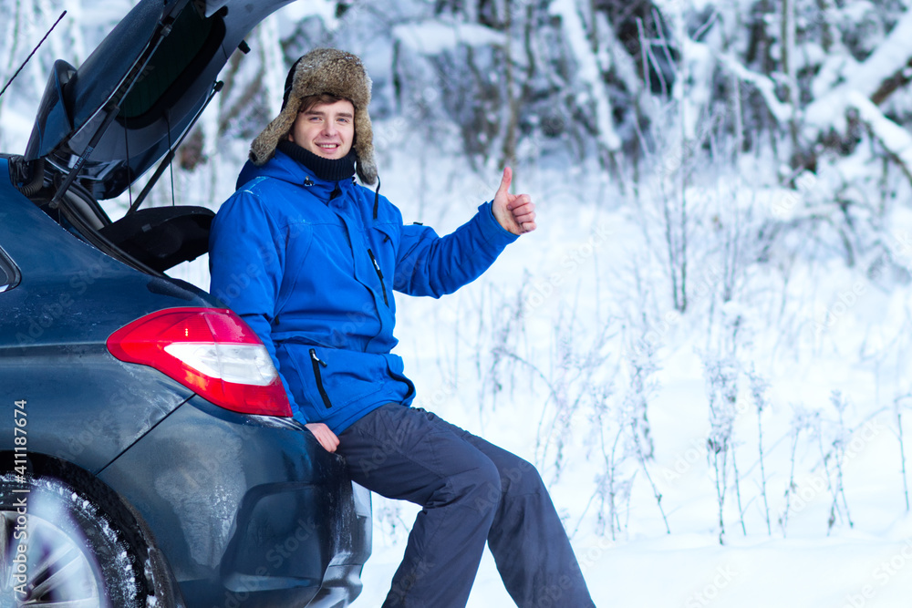 Handsome young happy man in warm winter clothes is sitting on a car trunk in a cold snowy day, smiling and showing thumb up. Travel by car Handsome young happy man in warm winter clothes is sitting on a car trunk in a cold snowy day, smiling and showing thumb up. Travel by car