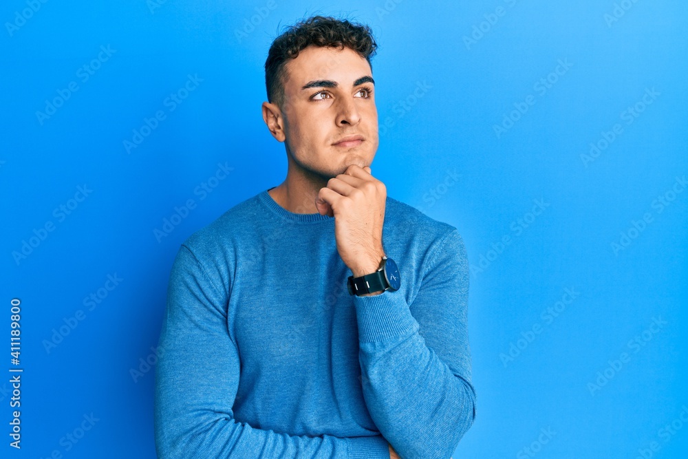 Hispanic young man wearing casual winter sweater with hand on chin thinking about question, pensive expression. smiling with thoughtful face. doubt concept. Hispanic young man wearing casual winter sweater with hand on chin thinking about question, pensive expression. smiling with thoughtful face. doubt concept.