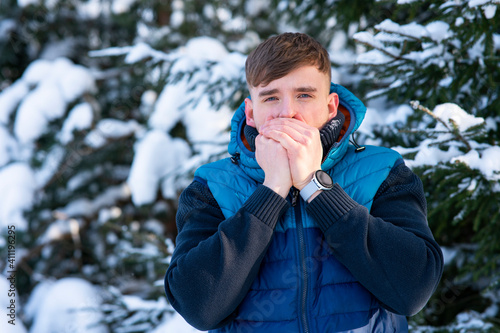 Young frozen guy is trying to warm his hands, breathes on his hands at a cold winter snowy forest