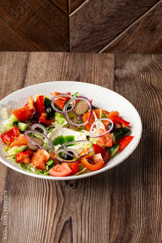 A tasty Greek salad with fresh vegetables: tomato, cucumber, red bel pepper, lettuce, onion, olives and cheese. Close-up on a white round plate on a wooden background. Healthy food.