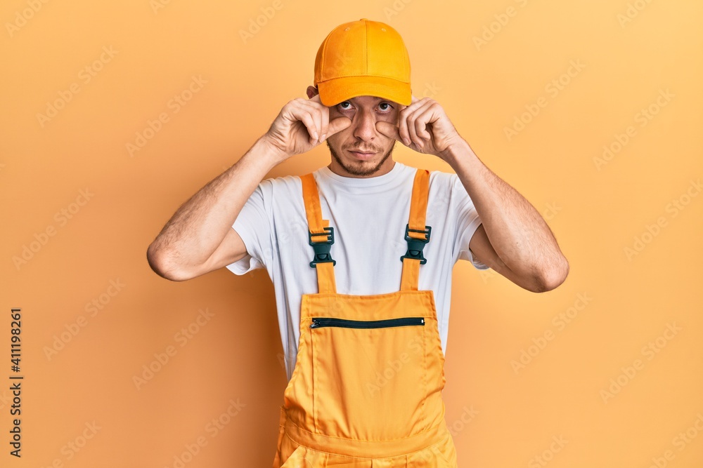 Hispanic young man wearing handyman uniform trying to open eyes with fingers, sleepy and tired for morning fatigue