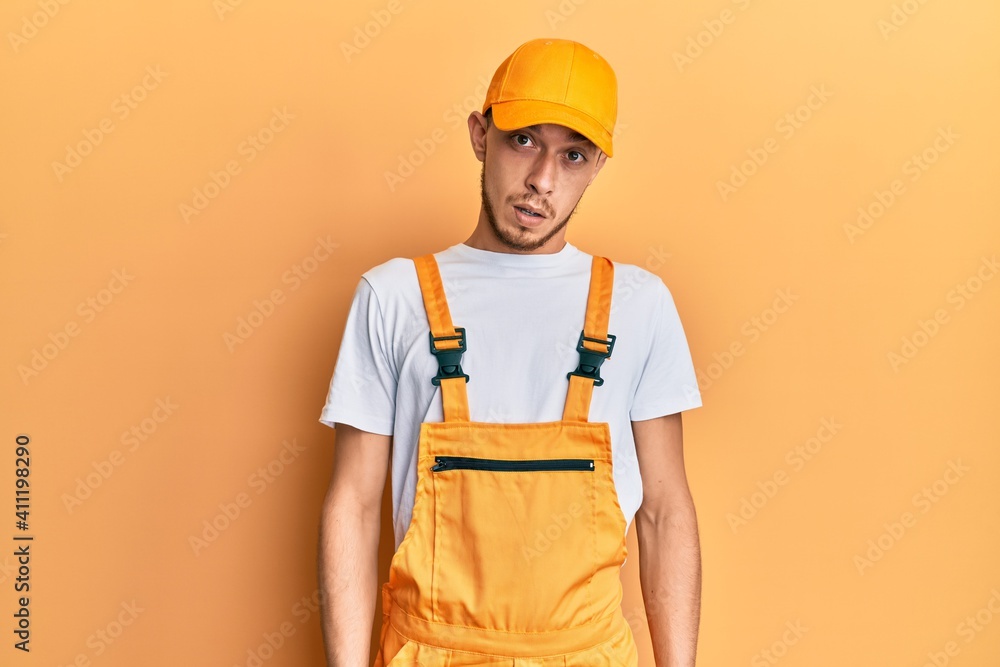 Hispanic young man wearing handyman uniform in shock face, looking skeptical and sarcastic, surprised with open mouth Hispanic young man wearing handyman uniform in shock face, looking skeptical and sarcastic, surprised with open mouth