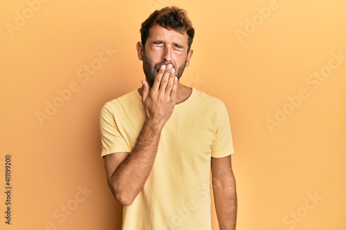 Handsome man with beard wearing casual yellow tshirt over yellow background bored yawning tired covering mouth with hand. restless and sleepiness.