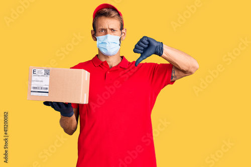 Handsome blond man with beard holding delivery box wearing medical mask with angry face, negative sign showing dislike with thumbs down, rejection concept