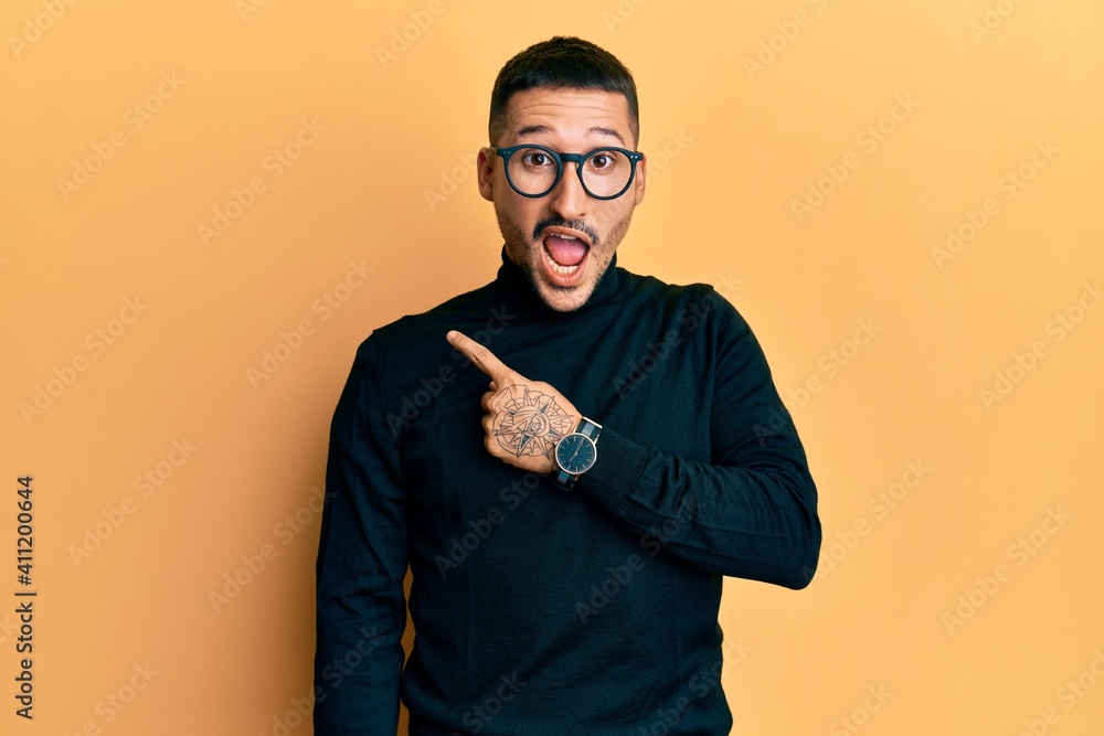 Handsome man with tattoos wearing turtleneck sweater and glasses surprised pointing with finger to the side, open mouth amazed expression. Handsome man with tattoos wearing turtleneck sweater and glasses surprised pointing with finger to the side, open mouth amazed expression.