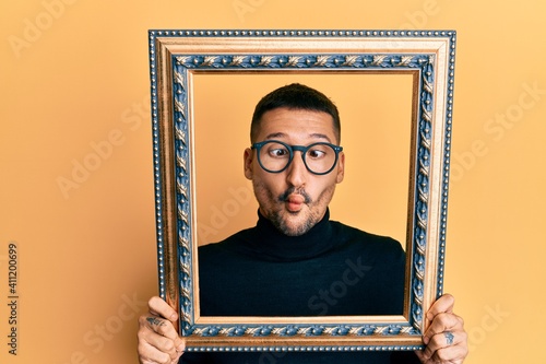 Handsome man with tattoos holding empty frame making fish face with mouth and squinting eyes, crazy and comical.