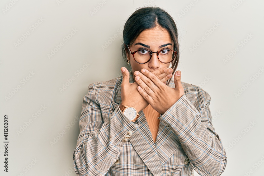 Young brunette woman wearing business jacket and glasses shocked covering mouth with hands for mistake. secret concept. Young brunette woman wearing business jacket and glasses shocked covering mouth with hands for mistake. secret concept.