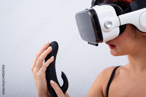 Caucasian red-haired woman wearing virtual reality glasses holding black dildo and looking lustfully. photo