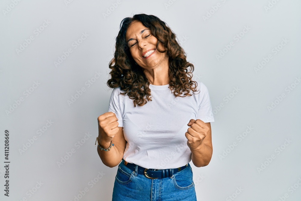 Middle age hispanic woman wearing casual white t shirt very happy and excited doing winner gesture with arms raised, smiling and screaming for success. celebration concept. Middle age hispanic woman wearing casual white t shirt very happy and excited doing winner gesture with arms raised, smiling and screaming for success. celebration concept.