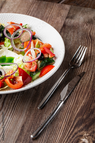 Greek salad with fresh vegetables  tomato  cucumber  red bel pepper  lettuce  onion  olives and cheese. Close-up on a white round plate with cutlery by side on a wooden background. Healthy food.