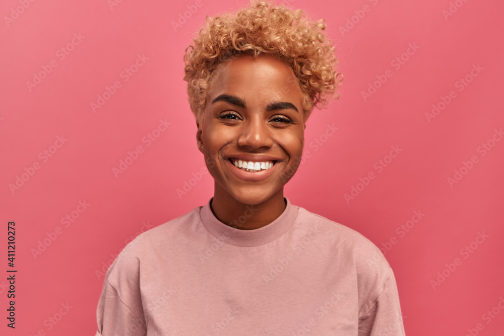 Studio portrait of young dark skinned woman with blonde hair in good mood standing on pink background. Joyful woman posing in studio expressing happiness. Concept of happiness and positive emotions. Studio portrait of young dark skinned woman with blonde hair in good mood standing on pink background. Joyful woman posing in studio expressing happiness. Concept of happiness and positive emotions.