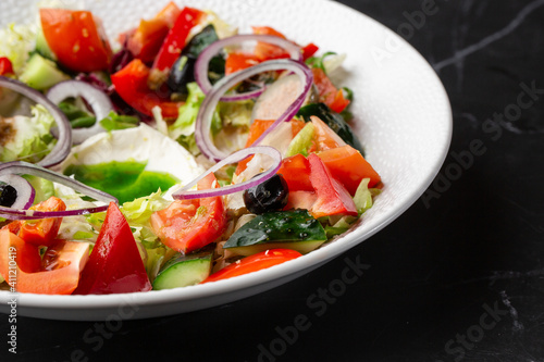 Greek salad with fresh vegetables: tomato, cucumber, red bel pepper, lettuce, red onion, olives and cheese. Macro close-up on a white round plateon a black marble background. Concept of healthy meal.