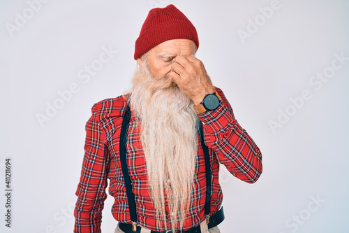 Old senior man with grey hair and long beard wearing hipster look with wool cap tired rubbing nose and eyes feeling fatigue and headache. stress and frustration concept.