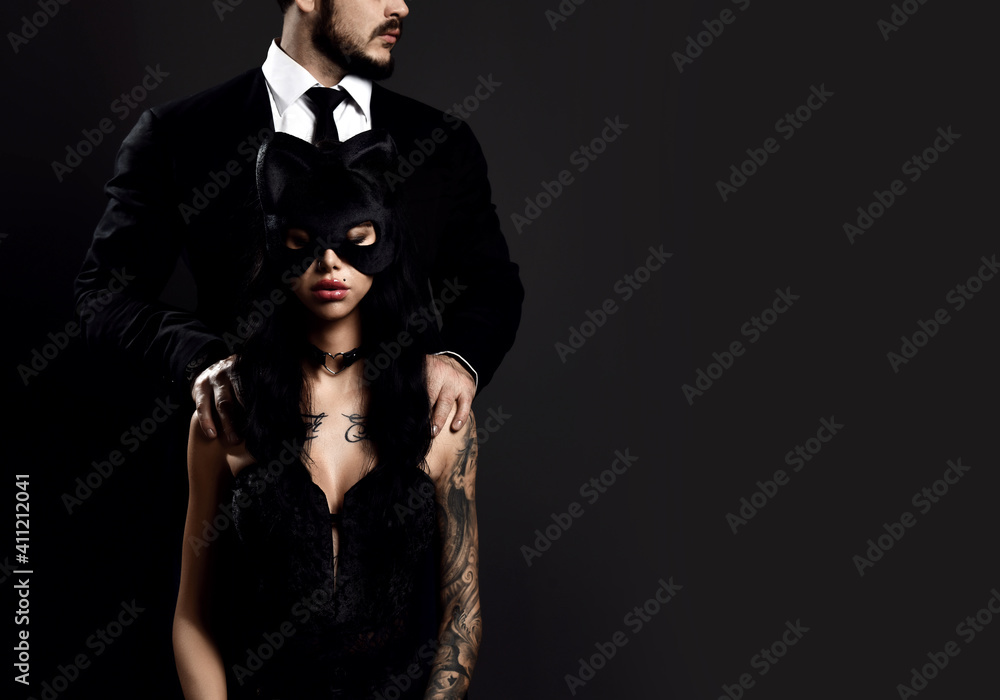 Pretty brunette woman with tattoo on body in black lingerie and sexy mask sitting with man in classical suit standing and hugging her from behind over dark background, copy space. Sexual games concept