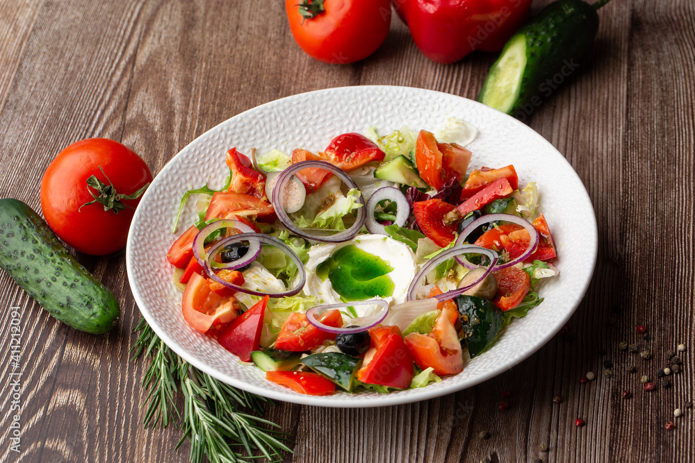 Greek salad with fresh vegetables: tomato, cucumber, red bel pepper, lettuce, onion, olives and cheese. Close-up on a white round plate on a wooden background. Salad menu with ingredients.