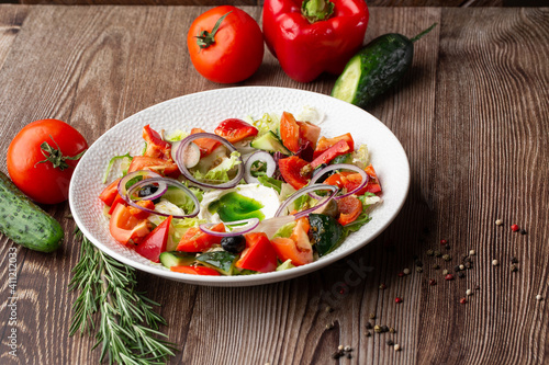 Greek salad with fresh vegetables: tomato, cucumber, red bel pepper, lettuce, onion, olives and cheese. Close-up on a white round plate on a wooden background. Salad menu with ingredients.