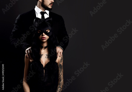 Obraz na płótnie Pretty brunette woman with tattoo on body in black lingerie and sexy mask sitting with man in classical suit standing and hugging her from behind over dark background, copy space. Sexual games concept w salonie