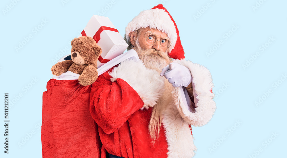 Obraz premium Old senior man with grey hair and long beard wearing santa claus costume holding bag with presents pointing with finger to the camera and to you, confident gesture looking serious