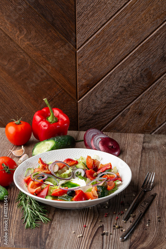 Greek salad with fresh vegetables: tomato, cucumber, red bel pepper, lettuce, onion, olives and cheese. Close-up on a white round plate on a wooden background. Salad menu with ingredients.