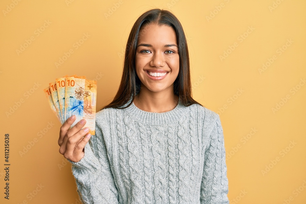 Beautiful hispanic woman holding 10 swiss franc banknotes looking positive and happy standing and smiling with a confident smile showing teeth Beautiful hispanic woman holding 10 swiss franc banknotes looking positive and happy standing and smiling with a confident smile showing teeth