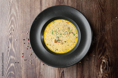 Yellow vegetable soup with onion, tomato, mushrooms, dill and parsley in a black bowl. Close-up top view on a wooden background with copy space. Healthy food menu. Delicious food.