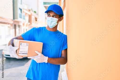 African delivery man wearing courier uniform outdoors wearing coronavirus safety mask holding cardboard parcel