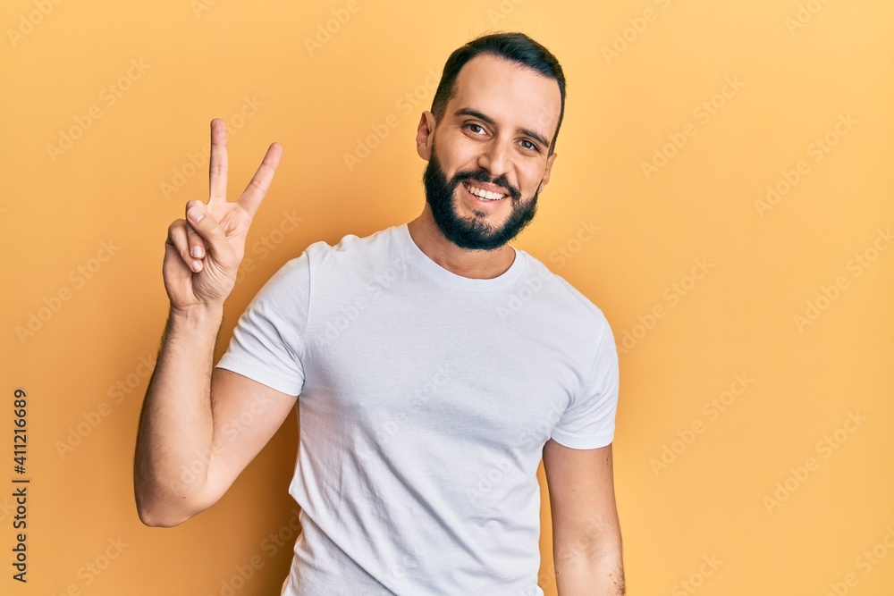 Young man with beard wearing casual white t shirt smiling with happy face winking at the camera doing victory sign. number two. Young man with beard wearing casual white t shirt smiling with happy face winking at the camera doing victory sign. number two.