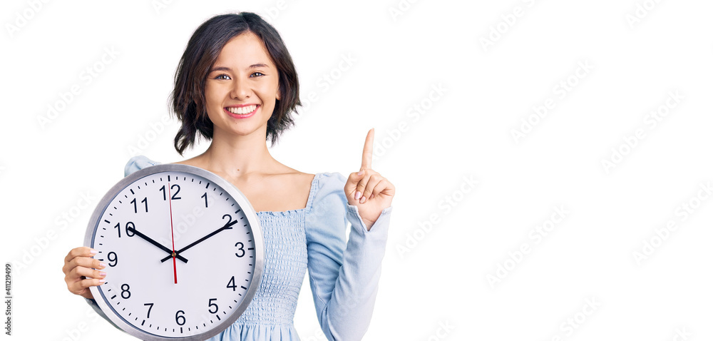Young beautiful girl holding big clock surprised with an idea or question pointing finger with happy face, number one Young beautiful girl holding big clock surprised with an idea or question pointing finger with happy face, number one