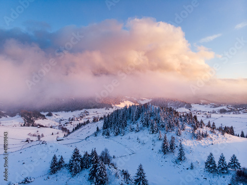 Rolling Clouds At Dense Spruce Coniferous Forest On Snowy White Hill Slope In Winter During Sunset. - Aerial Drone Shot. A forest Densely Covered With Fresh Snow