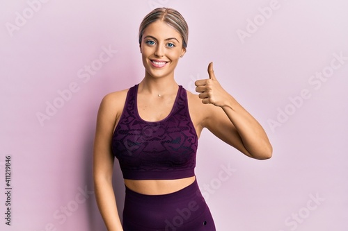 Beautiful blonde woman wearing sportswear over pink background doing happy thumbs up gesture with hand. approving expression looking at the camera showing success.