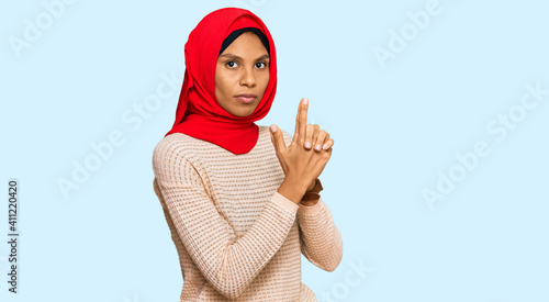 Young african american woman wearing traditional islamic hijab scarf holding symbolic gun with hand gesture, playing killing shooting weapons, angry face