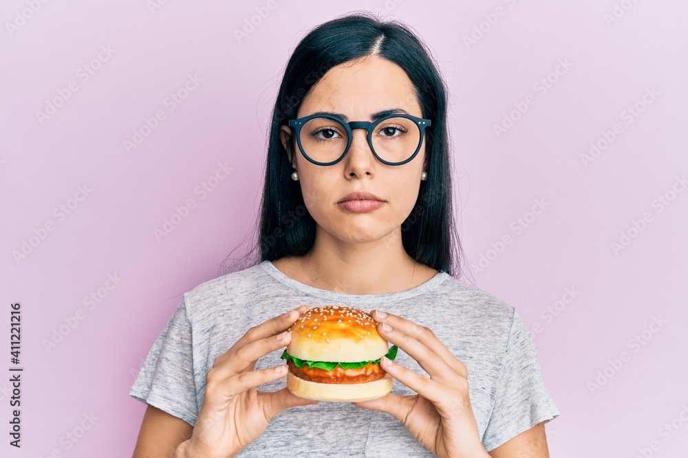 Beautiful young woman eating tasty hamburger relaxed with serious expression on face. simple and natural looking at the camera. Beautiful young woman eating tasty hamburger relaxed with serious expression on face. simple and natural looking at the camera.