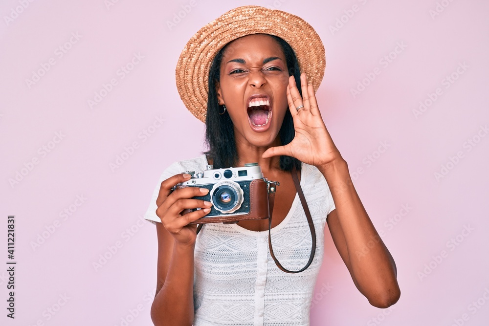 Young african american woman wearing summer hat holding vintage camera shouting and screaming loud to side with hand on mouth. communication concept.