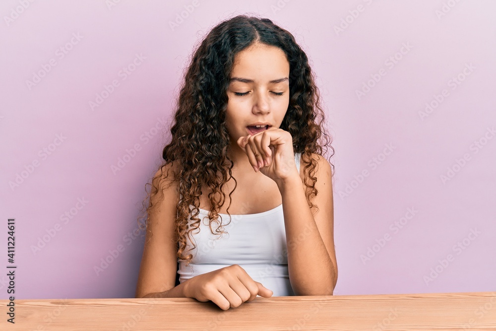 Teenager hispanic girl wearing casual clothes sitting on the table feeling unwell and coughing as symptom for cold or bronchitis. health care concept. Teenager hispanic girl wearing casual clothes sitting on the table feeling unwell and coughing as symptom for cold or bronchitis. health care concept.