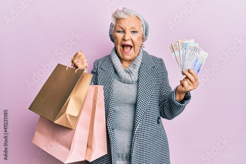 Senior grey-haired woman holding shopping bags and swedish krona banknotes smiling and laughing hard out loud because funny crazy joke.