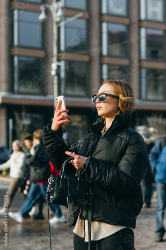 Attractive woman in sunglasses and fashionable clothes stands on the streets of a big city and takes photos on a smartphone camera.