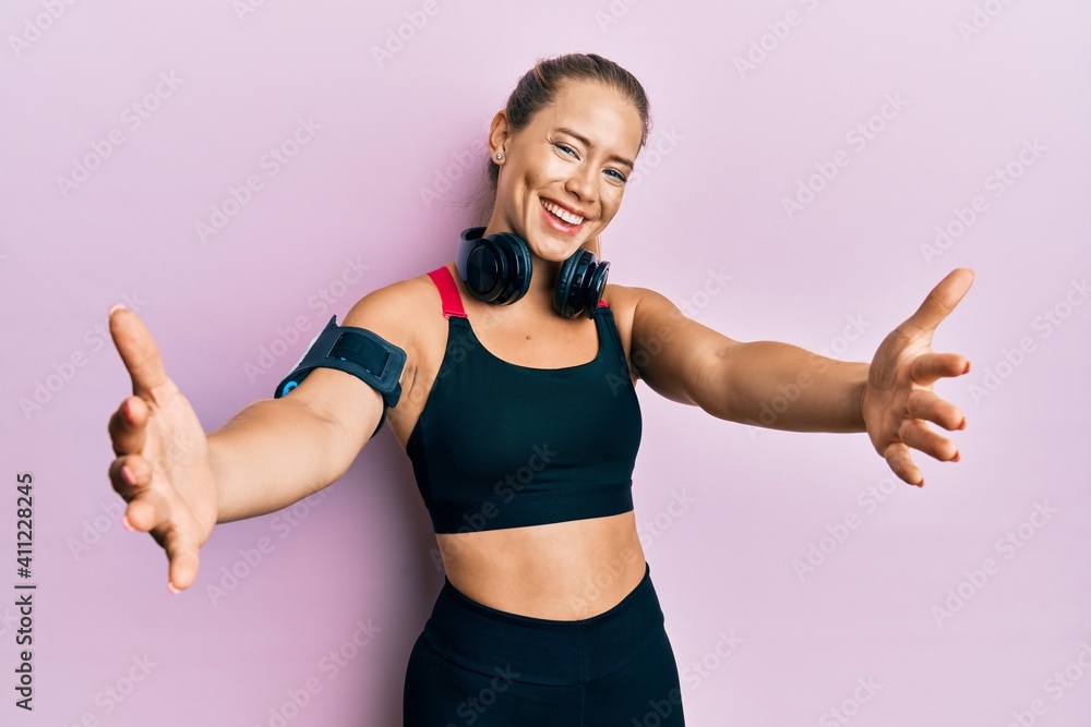 Beautiful young blonde woman wearing gym clothes and using headphones looking at the camera smiling with open arms for hug. cheerful expression embracing happiness. Beautiful young blonde woman wearing gym clothes and using headphones looking at the camera smiling with open arms for hug. cheerful expression embracing happiness.
