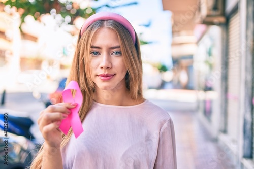 Young caucasian girl with serious expression holding pink breast cancer ribbon at the city.